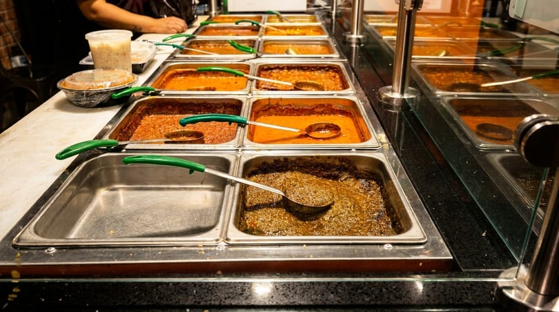 A buffet line with various Indian dishes in metal serving trays, including curries and stews.