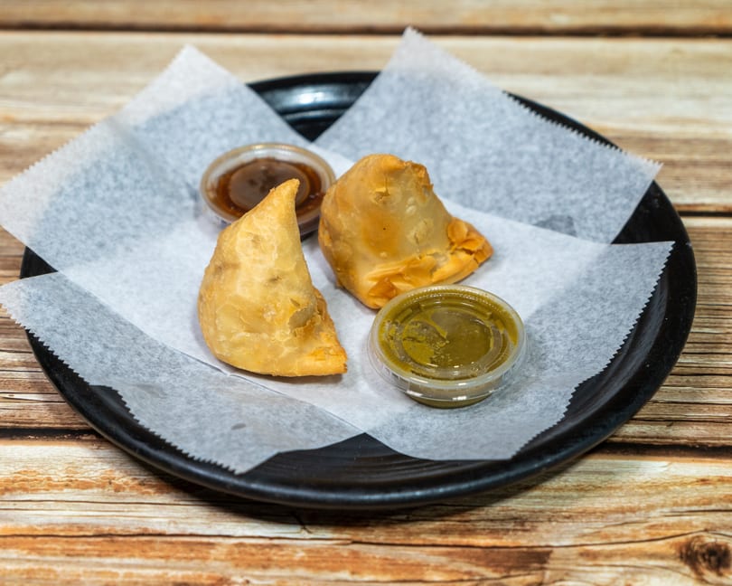 Two golden-brown samosas and two small containers of dipping sauces on a black plate.