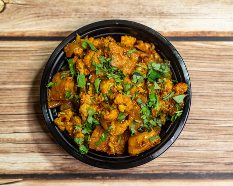 A black bowl filled with Aloo Gobi, a vibrant Indian dish of spiced potatoes and cauliflower, garnished with fresh cilantro.
