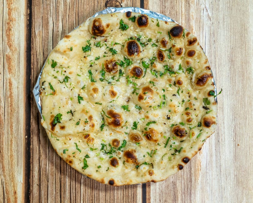 Garlic naan bread, topped with fresh green herbs, rests on a wooden table.