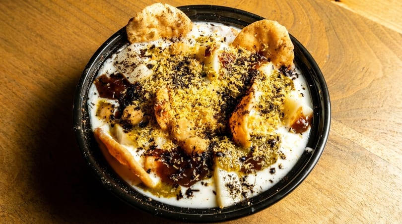 Close-up of a bowl of Dahi Bhalla, topped with yogurt, chutneys, and spices on a wooden table.