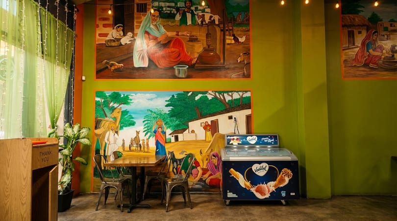 Restaurant interior with vibrant murals depicting rural life, a wooden table with chairs, and an ice cream freezer.