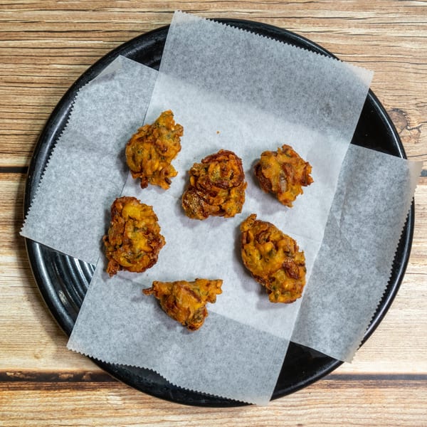 Six golden-brown pakoras, a fried Indian snack, on white parchment paper on a black plate.