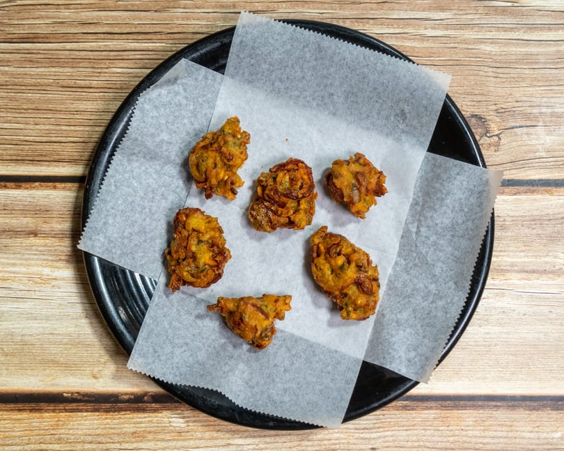 Six golden-brown pakoras, a fried Indian snack, on white parchment paper on a black plate.