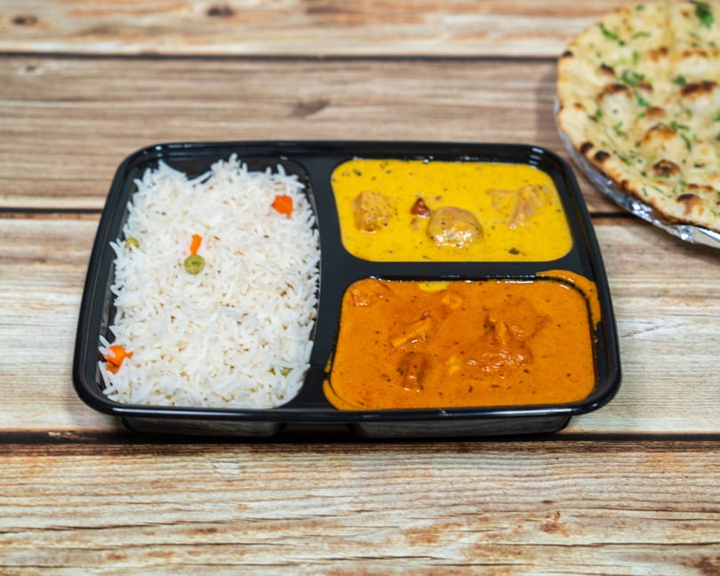 Indian takeout meal with white rice, two curries, and naan bread on a wooden table.