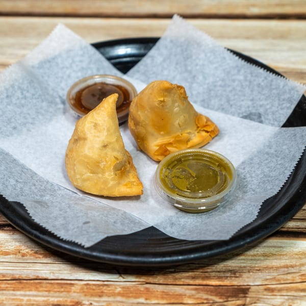 Two golden-brown samosas and two small containers of dipping sauces on a black plate with parchment paper.