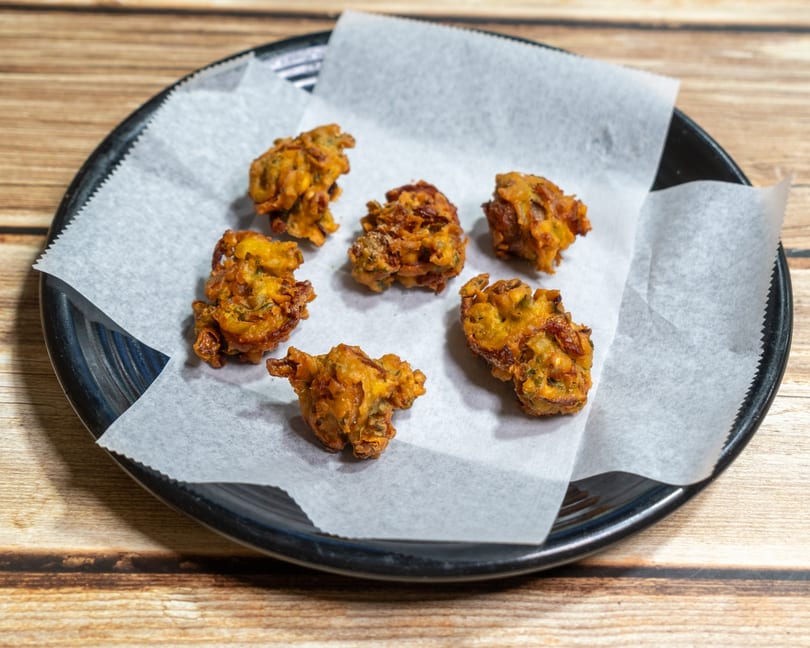 Six golden-brown pakoras, a fried Indian snack, on white parchment paper in a black bowl.