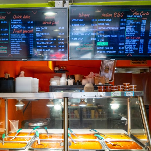 A food counter with hot food in metal trays and two digital menu boards above.