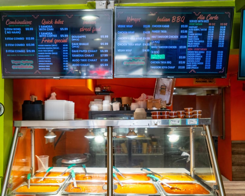 A food counter with hot food in metal trays and two digital menu boards above.