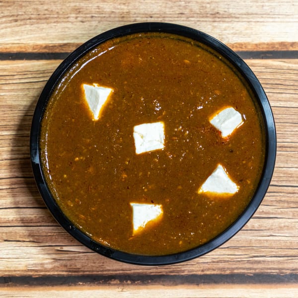Top-down view of a dark brown curry with white paneer cubes in a black bowl on a wooden table.