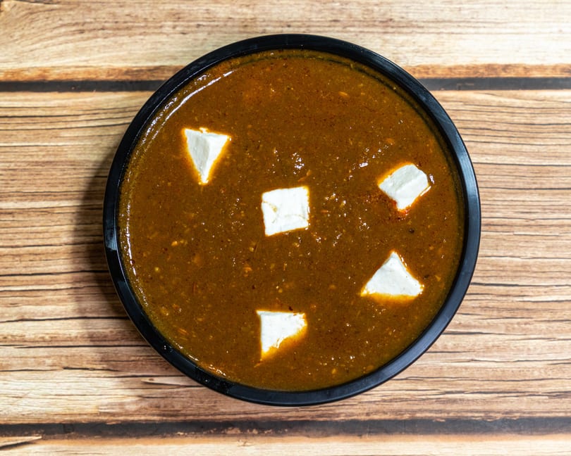 Top-down view of a dark brown curry with white paneer cubes in a black bowl on a wooden table.